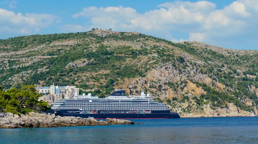 Large cruise ship docked near a rocky coastline with hills.